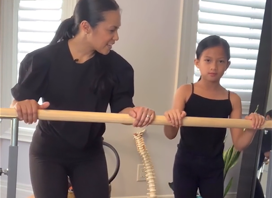 Older female dancer helping young girl at ballet bar with dance health better knee ankle alignment