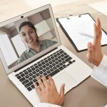 Female doctor gp consulting indian woman patient make online webcam video call on laptop screen. Telemedicine conference remote computer virtual visit in meeting. Over shoulder videocall view.